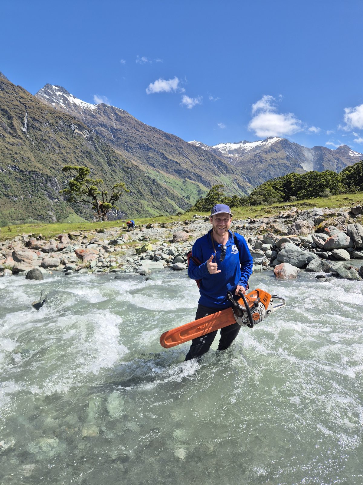 An arborist standing in a fast alpine river holding a chainsaw, snow-capped mountains behind