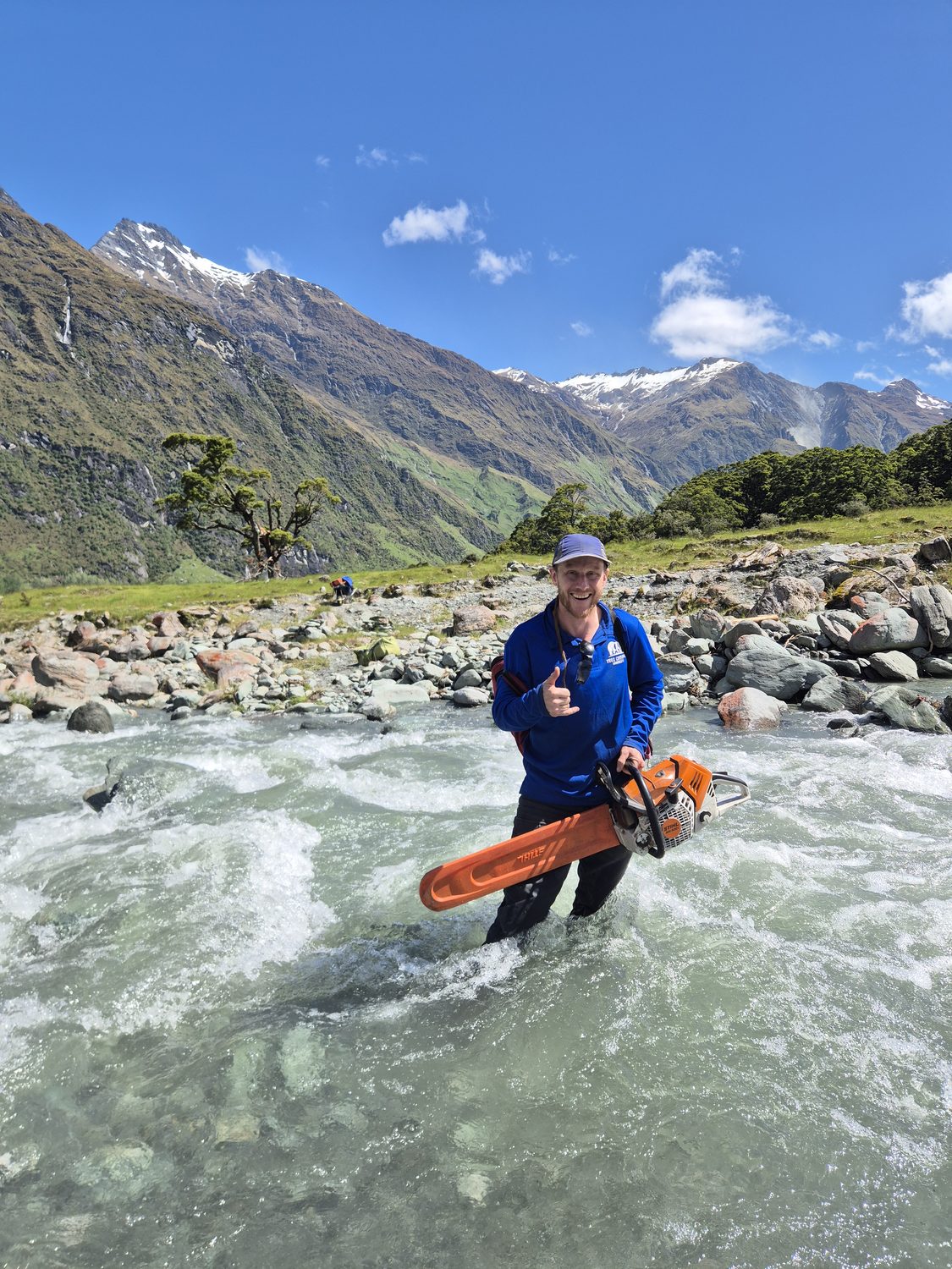 An arborist standing in a fast alpine river holding a chainsaw, snow-capped mountains behind