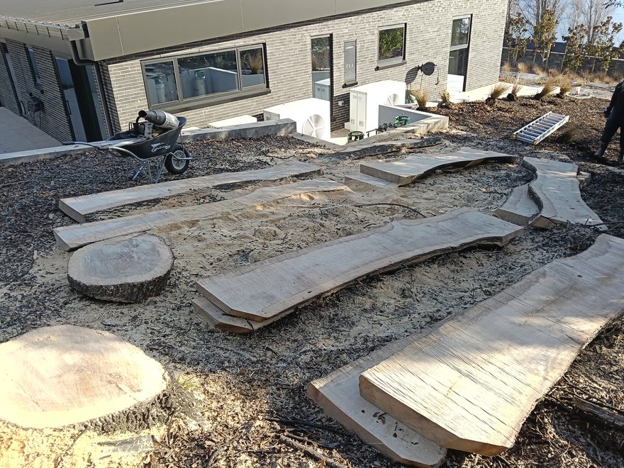 Milled timber slabs laid out to dry beside a new house, a wheelbarrow and a round of trunk alongside